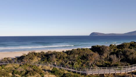Beach-waves-roll-in-as-tourists-walk-along-boardwalk-among-bush-landscape-coastline-during-evening-sunset-on-Bruny-Island-neck-isthmus-Tasmania,-Australia