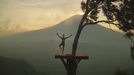 Fit-female-doing-tree-pose-on-view-platform-looking-at-Mount-Agung-with-mist,-sunset