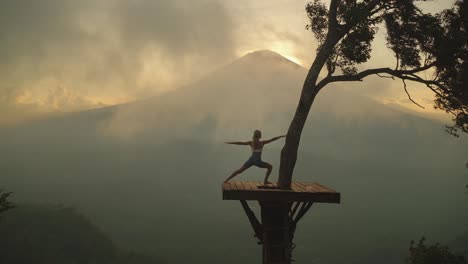 Woman-lunging-into-warrior-pose-on-tree-platform-with-moody-mist-and-view-of-mount-Agung