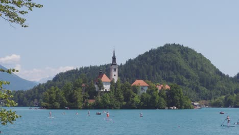 A-View-of-the-Bled-Island-in-Lake-Bled-from-the-Shore-with-Lots-of-Stand-Up-Paddle-Boarders-in-the-Foreground
