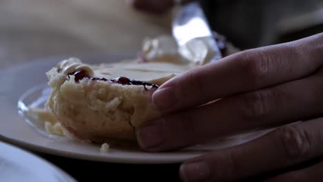 Slow-plan-around-female-hand-spreading-thick-cream-onto-jam-covered-traditional-bakes-scone-with-silver-metal-knife-on-white-plate-for-afternoon-tea