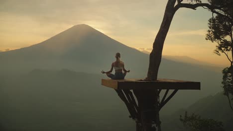 Blond-woman-in-yoga-pose-on-tree-hut-platform,-stunning-view-of-Mount-Agung,-sunset