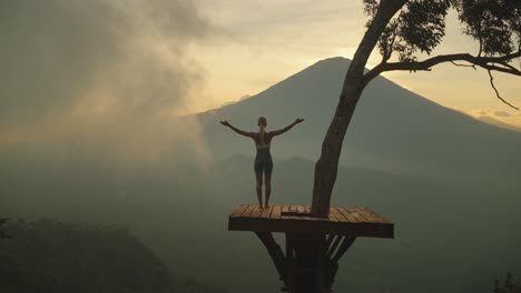 Slender-woman-on-wooden-tree-platform-raising-arms-into-greeting-pose,-mount-Agung