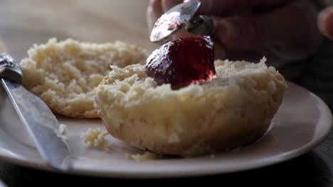 Hand-spoons-dollop-of-jam-onto-freshly-baked-steaming-traditional-scones-and-places-knife-on-white-plate-during-afternoon-tea