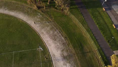 Aerial-tracking-shot-of-person-running-around-on-path-around-soccer-field-in-sunset---4K-top-down-of-athlete-during-training-session-outdoors