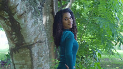 An-east-indian-model-with-curly-hair-stands-next-to-a-large-tree-trunk-in-green-dress