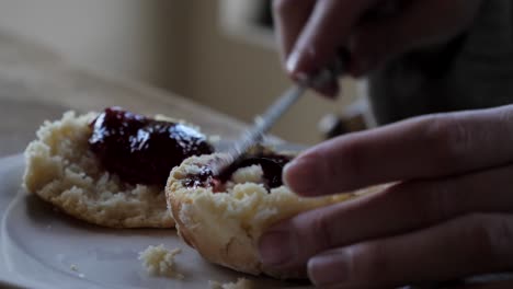 Up-close-shot-of-female-hand-spreading-dollop-of-jam-across-two-freshly-baked-traditional-homemade-scones-on-white-plate-and-timber-table-for-afternoon-tea