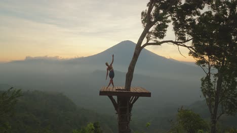 Woman-practicing-standing-side-stretch-yoga-pose-legs-crossed-on-view-platform-at-Mount-Agung,-sunset