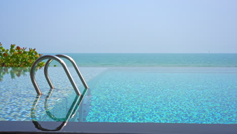 Crystal-clear-water-of-luxury-resort-infinity-swimming-pool-with-ladder-from-deep-to-shallow-and-blue-mosaic-tile-floor