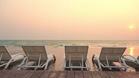 Empty-sun-loungers-positioned-in-the-shallow-incoming-tide-water-facing-a-colorful-sunset