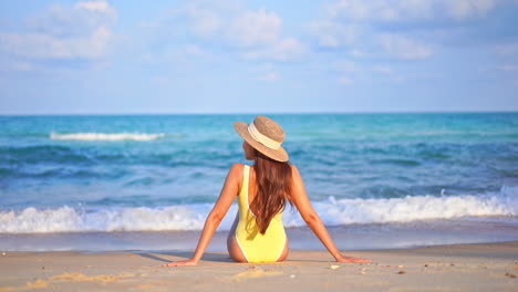 Back-view-of-woman-sitting-on-sandy-beach-with-yellow-swimsuit-and-hat