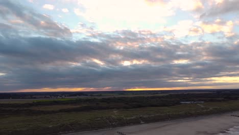 Aerial-shot-of-a-beautiful-cloudy-orange-sunset-skyline,-tracking-backwards-across-a-beach-and-deep-into-the-ocean,-Horsey-Gap,-Norfolk,-England