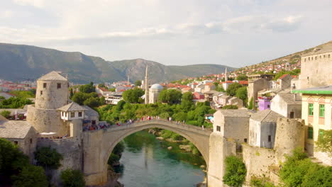 Drone-Ascend-Over-Neretva-River-With-People-Walking-At-Stari-Most,-Old-Bridge-In-Mostar,-Bosnia-And-Herzegovina