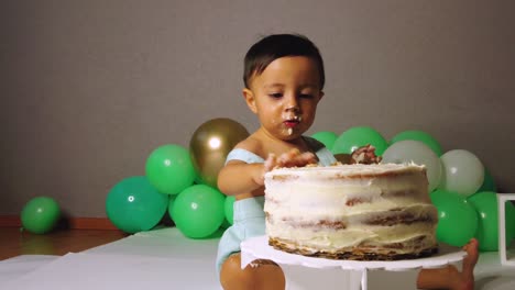 Süßer-Lateinamerikanischer-Junge,-Der-Seinen-1.-Geburtstag-Feiert-Und-Spaß-Daran-Hat,-Einen-Kuchen-Mit-Grünen-Luftballons-Im-Hintergrund-Zu-Beißen