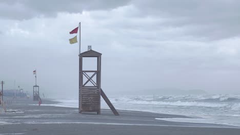 Ein-Leerer-Rettungsschwimmerturm-An-Einem-Traurigen-Grauen-Tag-Am-Strand,-Während-Der-Wind-Die-Roten-Und-Gelben-Flaggen-Weht