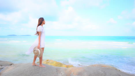 Woman-with-hat-in-hand-and-dressed-in-white-admiring-blue-sea-from-cliff
