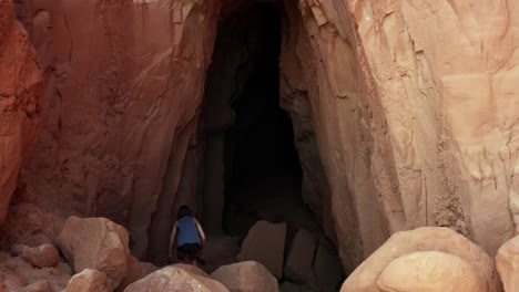 Aerial-drone-dolly-in-shot-of-a-slim-brown-haired-young-woman-climbing-down-red-rocks-and-walking-into-the-large-cave-entrance-of-the-Goblin's-Lair-inside-of-the-Utah-State-Park,-Goblin-Valley