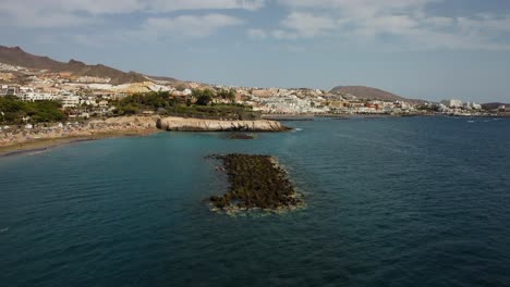 Impresionante-Toma-De-Drones-Del-Mar-Azul-En-La-Playa-Al-Sur-De-Tenerife-Los-Cristianos-En-El-Agua-De-La-Playa-Golpeando-Las-Rocas-Con-Un-Bonito-Paisaje-Con-Montañas-En-La-Parte-De-Atrás