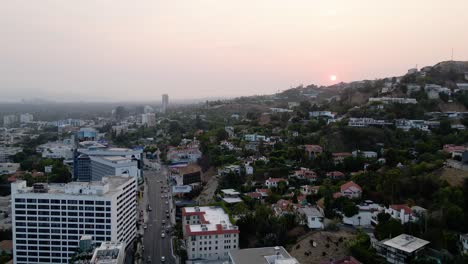 Aerial-view-of-a-sunset-above-a-hillside-neighborhood,-the-west-Hollywood-cityscape-and-the-Los-Angeles-skyline-background---pan,-drone-shot