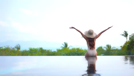 A-young-woman-in-a-one-piece-bathing-suit-and-sunhat-sits-on-the-invisible-edge-of-a-resort-swimming-pool-expresses-her-joy-by-throwing-her-arms-in-the-air