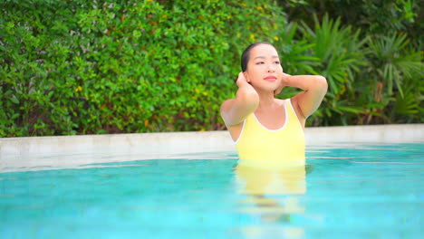 Sexy-sensual-woman-in-yellow-swimsuit-smooth-wet-hair-with-hands-inside-exotic-swimming-pool-in-Bali,-Indonesia,-slow-motion-portrait-front-view