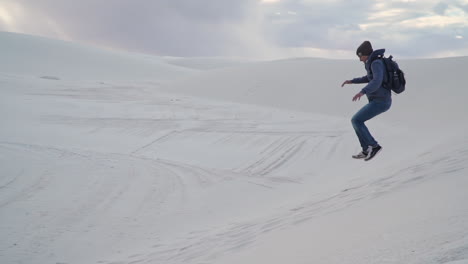 Young-man-playfully-runs-and-jumps-down-side-of-small-sand-dune,-slow-motion