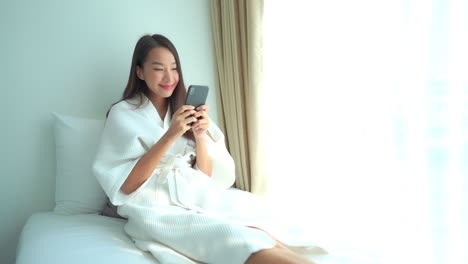 A-young-woman-in-a-terry-cloth-robe-smiles-as-sits-on-the-bed-and-reads-her-smartphone-screen