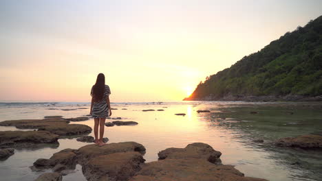 A-young-woman-with-her-back-to-the-camera-stands-on-the-exposed-rocks-from-the-low-tide-as-she-admires-the-colorful-sun-setting-behind-a-mountain