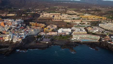 Beautiful-Drone-Shot-Seashore-Seaside-Of-Spain-Tenerife-Island-Volcano-In-the-Background-And-Mountains-Water-Sea-Blue-coast-Hotel-Pool-Buildings-Los-Gigantes