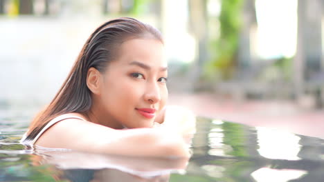 Dreaming-asian-woman-leaning-head-on-the-edge-on-infinity-swimming-pool-and-looking-at-camera-smiling-at-sunset-in-Thailand-resort,-face-closeup-slowmotion