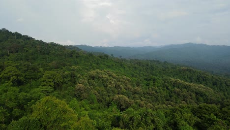 A-reverse-aerial-footage-of-this-rainforest,-beautiful-mountains-in-the-horizon,-rain-clouds-in-Thailand