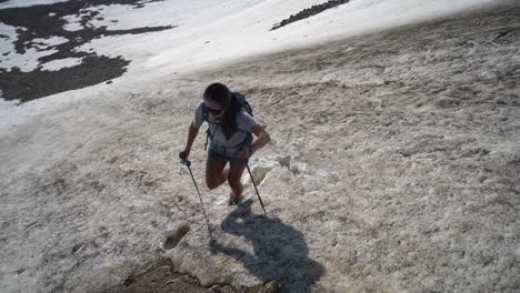 Woman-With-Trekking-Poles-Walking-Uphill-on-Snow-in-Mountain-Landscape-on-Sunny-Summer-Day,-Slow-Motion