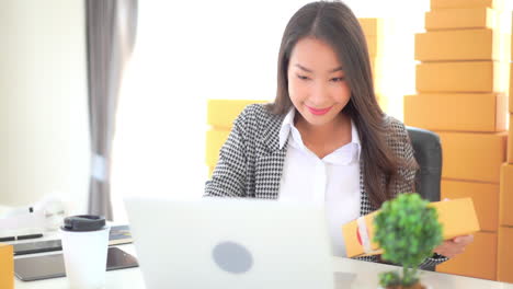 Asian-business-woman-entrepreneur-arranging-orders-while-sitting-at-the-work-desk-in-front-of-Laptop-with-yellow-parcel-in-hand,-yellow-boxes-on-background