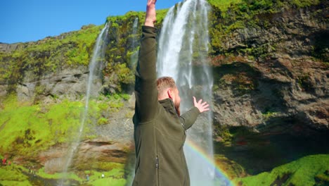 Man-Posing-Happily-For-The-Camera-With-A-Rainbow-On-The-Seljalandsfoss-Waterfall-In-South-Region-Of-Iceland
