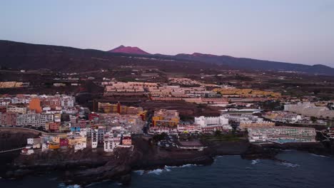 Paisaje-Agradable-Filmado-Con-Un-Dron-En-Un-Paisaje-Urbano-De-4k-En-El-Volcán-Costero-Junto-Al-Mar-Y-Las-Montañas-En-El-Mar-Trasero-Edificios-De-La-Hora-Dorada-De-La-Laguna-Azul-En-España-Tenerife-Los-Gigantes