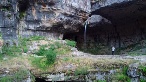 Waterfall-behind-a-natural-bridge-covered-in-lush-green-vegetation,-Baatara-Gorge-Waterfall-In-Lebanon---drone-ascending