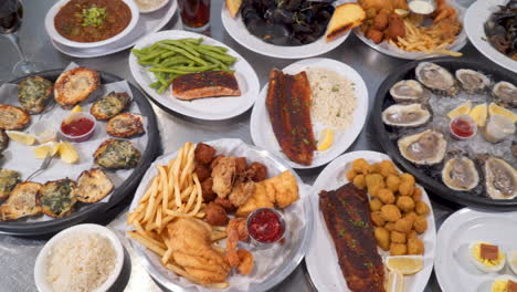 Birds-eye-view-of-restaurant-table-full-of-various-seafood-dishes-from-oysters-on-the-half-shell-to-blackened-fish-to-fried-shrimp-to-steamed-mussels,-over-top-slider-4K