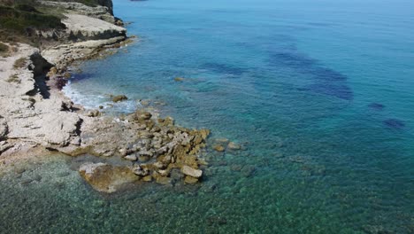 Aerial-view-of-rocks-and-clear-blue-sea-revealing-harbour-entrance,-clear-sunny-day-San-Stefonos-Corfu-Greece