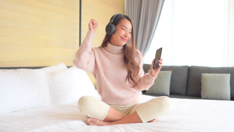 Smiling-Asian-girl-sitting-on-bed-listens-music-with-headphones