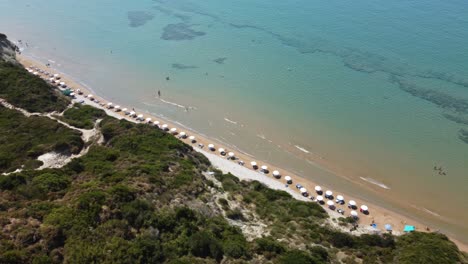 Aerial-view-of-a-mediterranean-beach-with-a-clear-turquoise-sea,-bathers,-umbrellas-and-tourists-during-Summertime-Corfu-Greece