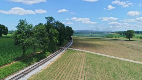 An-Aerial-View-of-a-Steam-Engine-Puffing-Smoke-and-Steam-with-Passenger-Coaches-Traveling-on-a-Single-Track-Turn-a-Curve-Thru-Trees-and-Farmland-Countryside-on-a-Beautiful-Cloudless-Spring-Day