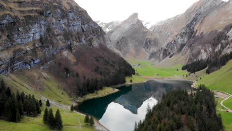 Aerial-flyover-alongside-Seealpsee-in-Appenzell,-Switzerland-revealing-a-reflection-of-the-Alpstein-peaks-on-the-lake-behind-the-trees