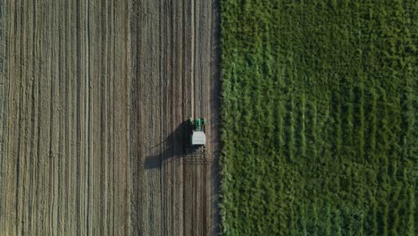 Aerial-birds-eye-shot-of-tractor-plowing-farm-field-beside-green-grass-field-in-sunlight