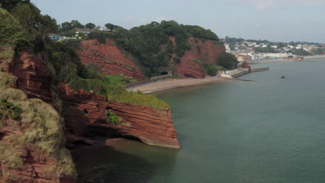 Aerial-shot-flying-along-the-Devon-coast-with-red-cliffs-and-sea-caves-with-Dawlish-town-in-the-distance-on-a-sunny-day