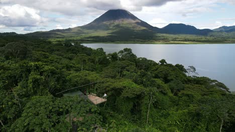 Aerial-view-Ascending-shot,-a-woman-standing-on-a-view-deck,-scenic-view-of-lake-in-Costa-Rica,-Arena-Volcano-in-the-background