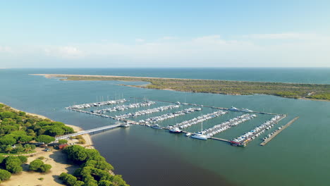 Aerial-pull-back-from-Puerto-Marina-El-Rompido-docks-with-many-yachts,-sailboats,-watercraft,-and-pier-on-Piedras-river,-Spain,-Atlantic-Ocean-on-the-background-behind-the-spit