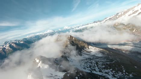 FPV-Drone-Aerial-Shot-above-clouds-over-steep-mountain-in-summer,-in-Switzerland,-with-partly-cloudy-sky-and-sun-and-cloud-inversion,-close-to-mountain-glacier