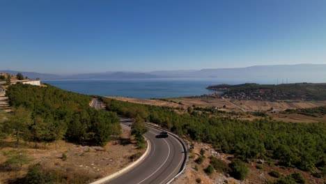 Car-drives-on-panoramic-winding-road-near-blue-lake-and-panoramic-coast-of-touristic-village-in-Albania