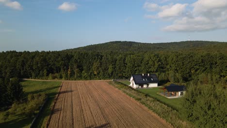 Aerial-backwards-shot-of-farmland-field-with-luxury-house-and-solar-panels-on-roof-in-rural-suburb-of-Poland---Beautiful-green-forest-trees-in-background