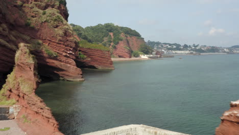 Aerial-drone-shot-flying-through-the-rocks-in-Dawlish,-along-the-Devon-coast-with-red-cliffs-and-sea-caves-with-calm-water-on-a-sunny-day